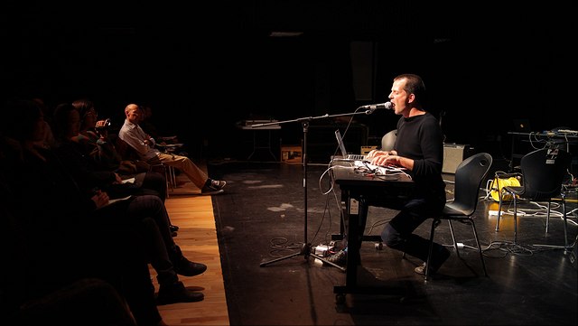 Alessandro Bosetti sitzt während seiner Sound Poetry Performance an einem Tisch mit Sampler und Laptop, spricht in ein Mikrofon, links ist ein Teil des Publikums zu sehen.