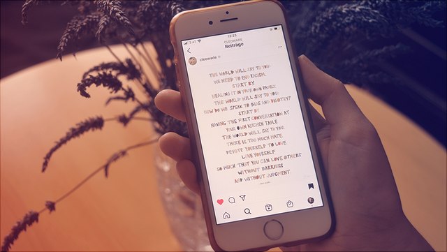 Im Bild eine Hand, die ein Smartphone hält, in dem das in bunten Großbuchstaben verfasste Instapoem von Cleo Wade zu lesen ist. Hinter dem Smartphone ein Lavendelstrauß auf einem Tisch.